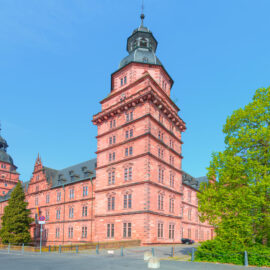 Das Bild zeigt Schloss Johannisburg in Aschaffenburg, ein historisches Gebäude aus rotem Backstein mit mehreren Türmen und schwarzem Dach, umgeben von Bäumen und Grünflächen.