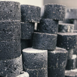 Das Bild zeigt mehrere zylindrische Kernbohrproben aus Asphalt, die aus Straßen entnommen wurden. Die Proben sind in mehreren Stapeln angeordnet und weisen eine raue Oberfläche sowie unterschiedliche Grautöne bis Schwarz auf.