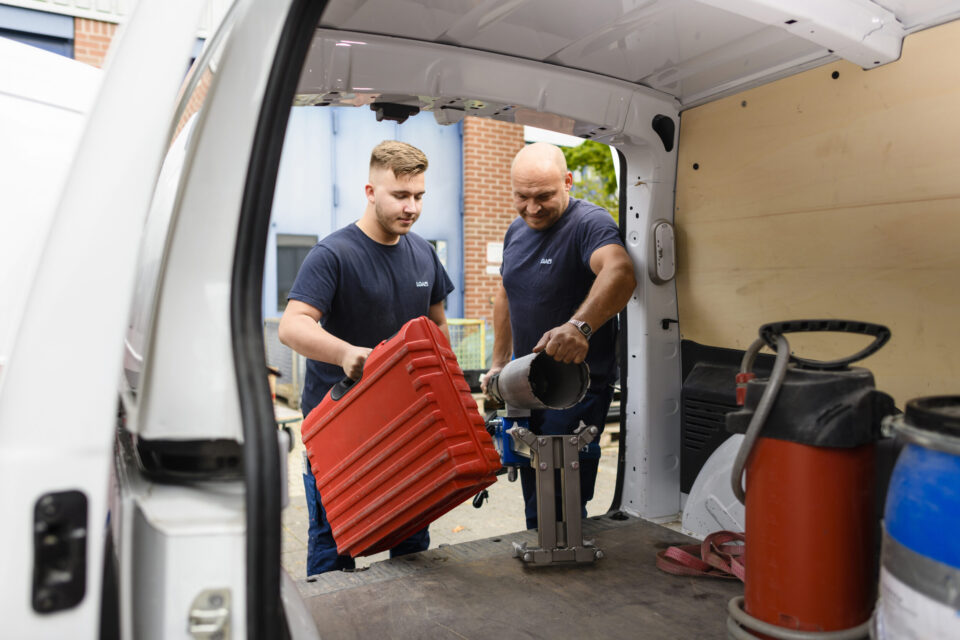 Marco Mehrer (links) und Daniel Schumann (rechts) beim einladen von Geräten und einem großen roten Werkzeugkoffer in einen Kastenwagen.