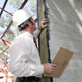 Das Bild zeigt eine Person mit weißem Schutzhelm und hellem Hemd, die in der linken Hand ein Klemmbrett hält. Die Person inspiziert oder arbeitet an der Kante einer großen Wand, die sich im Bau befindet. Im Hintergrund sind Metallkonstruktionen und Bäume zu sehen, was auf eine Baustelle im Freien hinweist.
