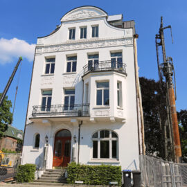 Das Bild zeigt ein weißes, mehrstöckiges Gebäude mit aufwendigen architektonischen Details. Mehrere Balkone mit schwarzen Geländern und große Fenster prägen die Fassade. Auf der linken Seite befinden sich Baumaschinen, darunter ein Kran und eine Bohrvorrichtung.