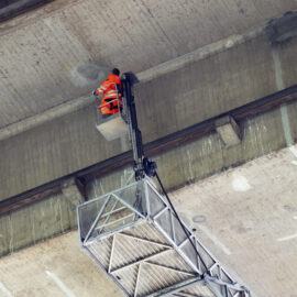 Das Bild zeigt eine Person in einem orangefarbenen Schutzanzug, die auf einer angehobenen Arbeitsplattform steht und an die Unterseite eines großen Betonbauwerks, einer Brücke inspiziert. Die Plattform ist an einem ausfahrbaren Arm befestigt, der den Zugang zu schwer erreichbaren Stellen ermöglicht. Die Betonoberfläche weist Gebrauchsspuren und Markierungen auf.