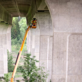 Das Bild zeigt eine große Betonbrücke mit mehreren Pfeilern. Eine Person befindet sich in einem orangefarbenen Arbeitskorb, der an einem langen gelben Ausleger befestigt ist, und arbeitet an der Unterseite der Brücke. Die Umgebung ist von grünen Bäumen und Vegetation geprägt.