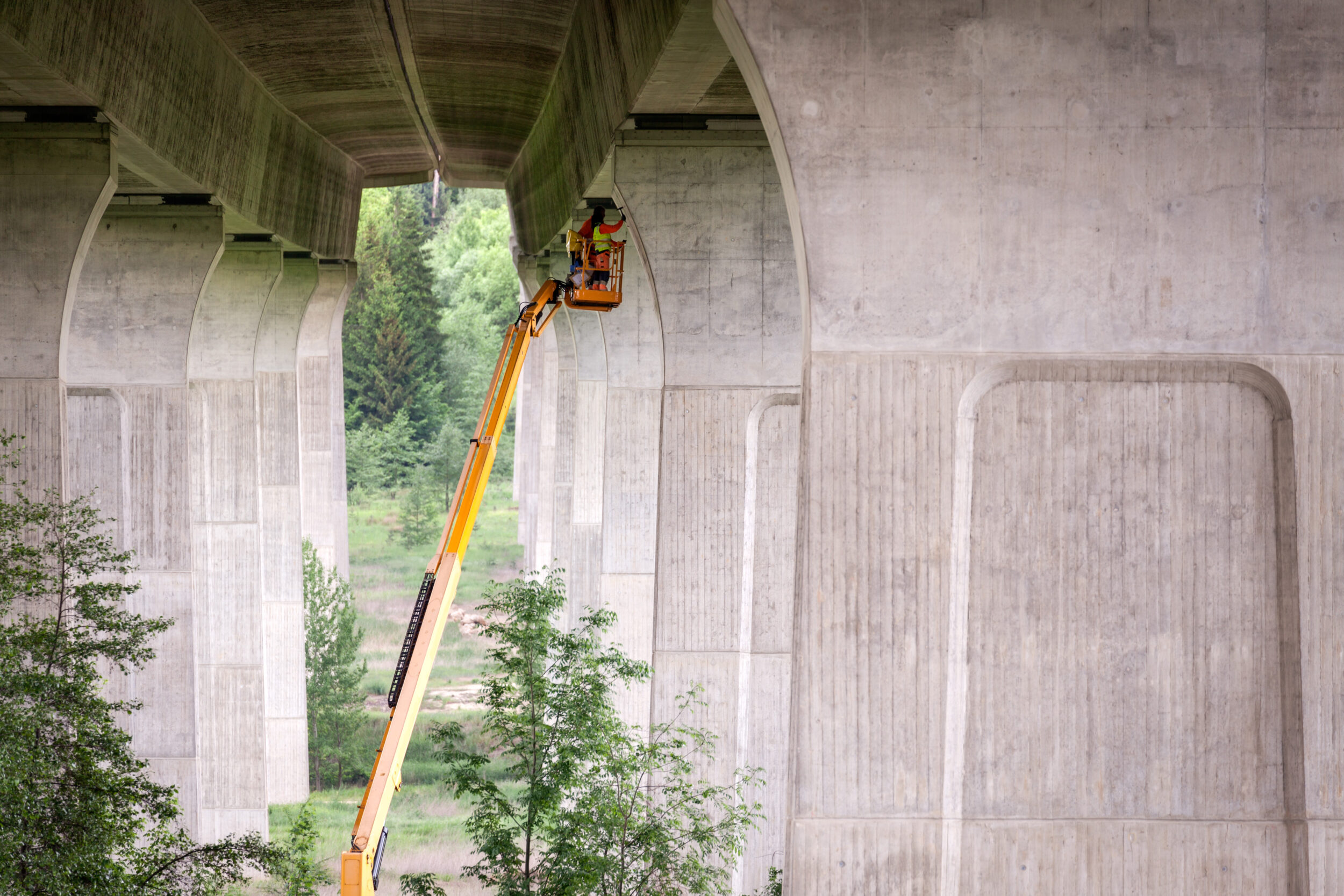 Das Bild zeigt eine große Betonbrücke mit mehreren Pfeilern. Eine Person befindet sich in einem orangefarbenen Arbeitskorb, der an einem langen gelben Ausleger befestigt ist, und arbeitet an der Unterseite der Brücke. Die Umgebung ist von grünen Bäumen und Vegetation geprägt.