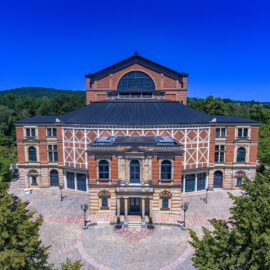 Das Bild zeigt das Bayreuther Festspielhaus mit seiner markanten Fassade, dreieckigem Giebel und Rundbogenfenstern, umgeben von Kopfsteinpflaster und Bäumen in parkähnlicher Umgebung.
