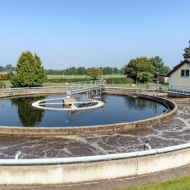 Das Bild zeigt ein rundes Betonbecken einer Kläranlage, ein Absetz- oder Klärbecken. In der Mitte befindet sich eine mechanische Vorrichtung zur Entfernung von Feststoffen aus dem Wasser. Im Hintergrund steht ein kleines Gebäude mit Solarpaneelen auf dem Dach, umgeben von Bäumen und Grünflächen.