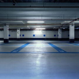 Das Bild zeigt eine leere Tiefgarage mit blauen und weißen Markierungen auf dem Boden zur Kennzeichnung der Parkplätze. Die Decke ist mit verschiedenen Rohrleitungen und Beleuchtungseinrichtungen ausgestattet, und mehrere Betonsäulen sind mit dem Buchstaben C gekennzeichnet. Die Beleuchtung ist künstlich und erzeugt eine leicht gedämpfte Atmosphäre.