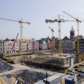 Das Bild zeigt eine Baustelle mit mehreren Kränen und teilweise errichteten Gebäuden. Die Umgebung besteht aus älteren Gebäuden, was auf eine städtische Lage hinweist. Im Hintergrund sind bunte Häuserreihen und ein Kirchturm mit Spitze zu sehen.
