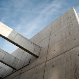Das Bild zeigt eine Nahaufnahme eines Betonbauwerks mit großen, ebenen Flächen und auskragenden Trägern. Im Hintergrund ist der Himmel zu sehen. Die Struktur wirkt massiv und technisch.
