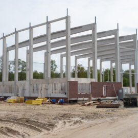 Das Bild zeigt die Baustelle einer neuen Produktionshalle mit einem Skelett aus Stahlbeton. Im unteren Bereich ist eine Verblendung aus Mauerwerk zu sehen. Auf dem Gelände liegen verschiedene Baumaterialien und Baugeräte.