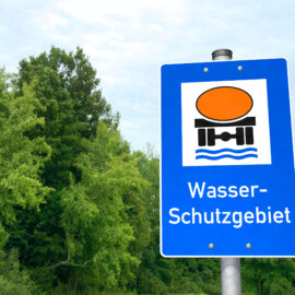 Das Bild zeigt ein blaues Hinweisschild mit einem weißen Quadrat, in dem ein orangefarbener Kreis, schwarze Formen und blaue Wellenlinien abgebildet sind, die Wasser und Infrastruktur symbolisieren. Unter der Grafik steht der Schriftzug Wasser-Schutzgebiet. Im Hintergrund sind grüne Bäume und Pflanzen zu sehen.
