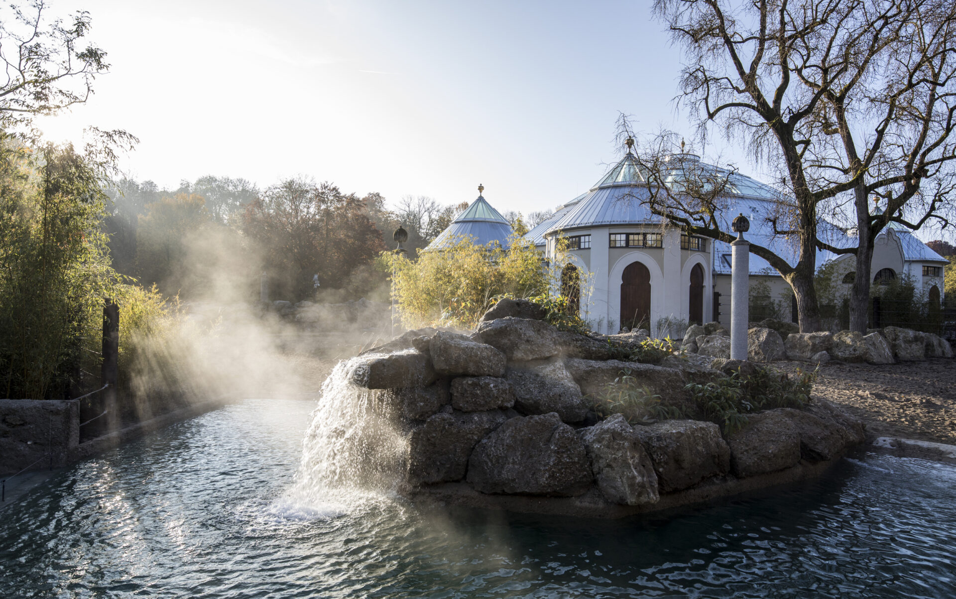 Das Bild zeigt eine ruhige Außenaufnahme mit einem kleinen Wasserfall, der über Felsen in ein Wasserbecken fließt. Im Hintergrund stehen weiße Gebäude mit blauen Dächern und Rundbögen, umgeben von Bäumen und Pflanzen. Ein leichter Nebel liegt über der Szene, vermutlich durch die Gischt des Wassers.