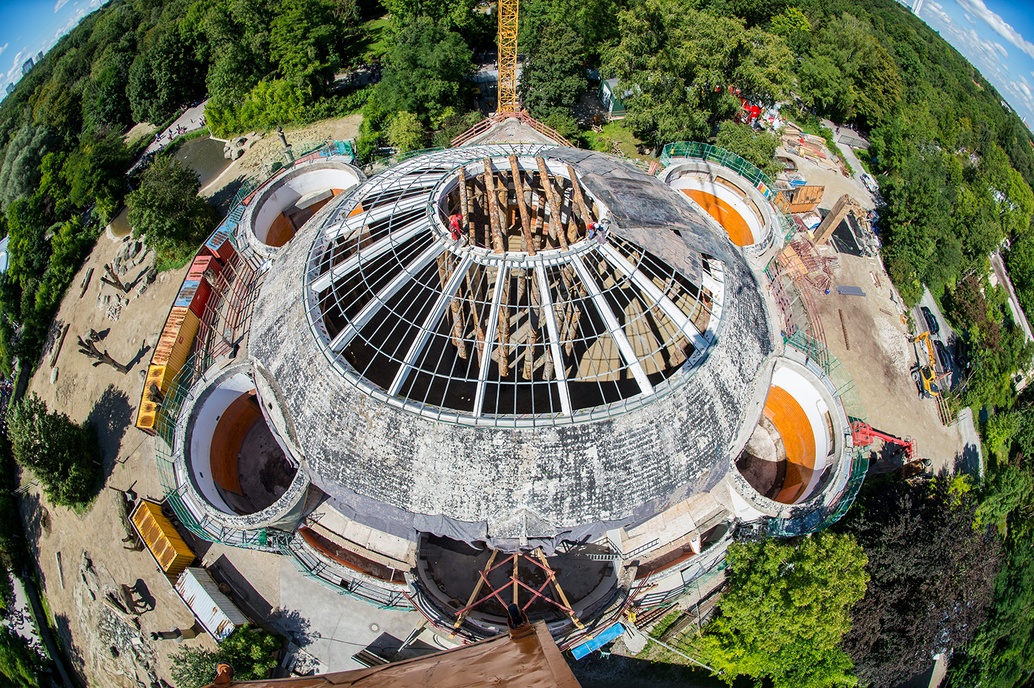 Das Bild zeigt eine Luftaufnahme einer Baustelle mit einem großen, runden Gebäude, das sich im Umbau oder Bau befindet. Die zentrale Kuppelstruktur ist von Gerüsten und freiliegenden Trägern umgeben, daneben befinden sich mehrere kleinere runde Bauwerke. Um das Gelände herum sind Bäume und Grünflächen zu sehen.