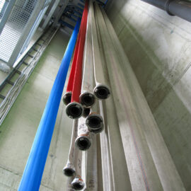 Das Bild zeigt mehrere lange Schläuche, die senkrecht in einem Lagerbereich hängen. Die Schläuche sind in verschiedenen Farben wie Blau, Rot und Weiß zu sehen und unten an Metallkupplungen befestigt. Sie sind in parallelen Reihen angeordnet, im Hintergrund sind Betonwände und Metallstrukturen zu erkennen.