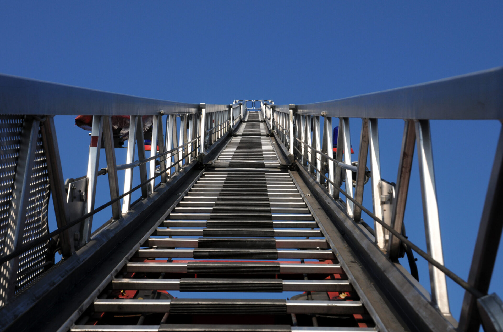 Das Bild zeigt den Blick nach oben entlang einer ausfahrbaren Metallleiter, eines Feuerwehrfahrzeugs. Die Leiter ragt steil in den klaren blauen Himmel, wobei sich die Seiten durch die Perspektive zur Bildmitte hin verjüngen.