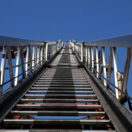 Das Bild zeigt den Blick nach oben entlang einer ausfahrbaren Metallleiter, eines Feuerwehrfahrzeugs. Die Leiter ragt steil in den klaren blauen Himmel, wobei sich die Seiten durch die Perspektive zur Bildmitte hin verjüngen.