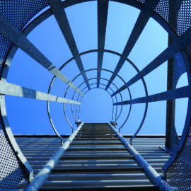 Das Bild zeigt den Blick nach oben entlang einer Feuerleiter, die von einem zylindrischen Schutzkäfig umgeben ist. Der Käfig besteht aus gelochten Metallplatten und horizontalen Streben, die ringförmig um die Leiter verlaufen. Im Hintergrund ist ein klarer blauer Himmel zu sehen.