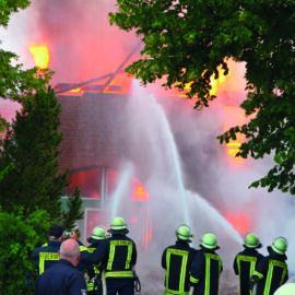 Das Bild zeigt mehrere Feuerwehrleute in Uniform, die ein brennendes Gebäude löschen. Flammen und Rauch sind vor allem am Dach und in den oberen Bereichen des Gebäudes sichtbar. Die Feuerwehrleute spritzen mit Schläuchen Wasser auf das Feuer, während Bäume und Grünflächen Teile des Gebäudes umgeben.