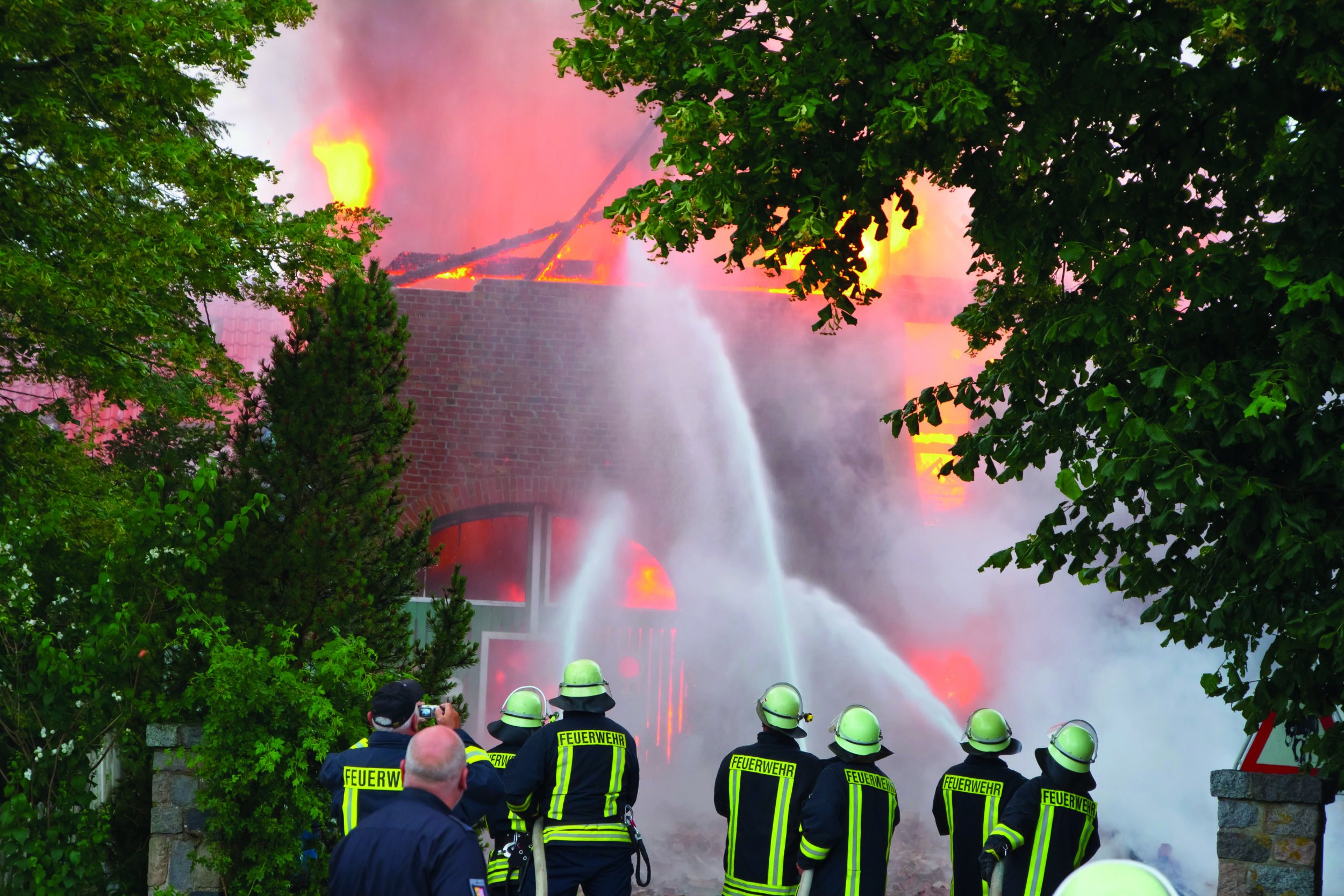 Das Bild zeigt mehrere Feuerwehrleute in Uniform, die ein brennendes Gebäude löschen. Flammen und Rauch sind vor allem am Dach und in den oberen Bereichen des Gebäudes sichtbar. Die Feuerwehrleute spritzen mit Schläuchen Wasser auf das Feuer, während Bäume und Grünflächen Teile des Gebäudes umgeben.