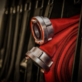 Das Bild zeigt eine Nahaufnahme eines roten Feuerwehrschlauchs mit Metallkupplungen, der ordentlich auf einem Gestell aufgerollt ist. Im Hintergrund befinden sich weitere ähnliche Schläuche und Ausrüstungsgegenstände, was auf eine Feuerwache oder eine Notfallausrüstung hinweist.
