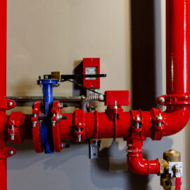 Das Bild zeigt einen Abschnitt eines Rohrleitungssystems mit mehreren roten Rohren und Verbindungsstücken. In der Mitte befindet sich ein blaues Ventil, das mit den roten Rohren verbunden ist. An einem der Rohre ist ein kleines Steuergerät angebracht.