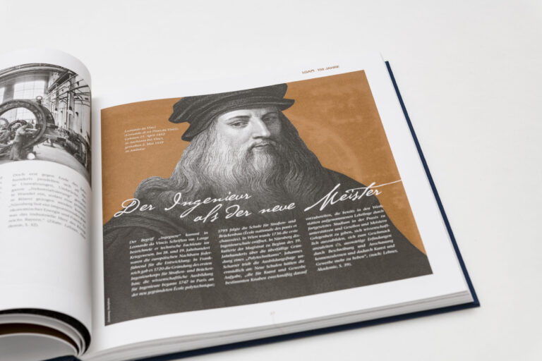 Das Bild zeigt ein aufgeschlagenes Buch mit einer Schwarz-Weiß-Fotografie von Industrieanlagen auf der linken Seite. Die rechte Seite enthält eine Illustration einer Person mit langem Haar und Hut sowie den Titel „Der Ingenieur als der neue Meister“.