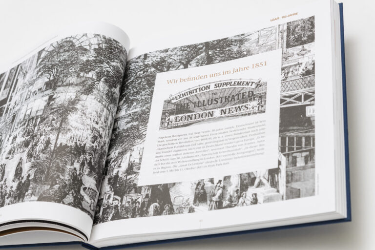 Das Bild zeigt ein aufgeschlagenes Buch mit einer doppelseitigen Schwarz-Weiß-Illustration, die eine historische Szene mit Menschen und Gebäuden darstellt. Auf der rechten Seite befindet sich ein Textfeld mit Informationen zur Großen Ausstellung von 1851 in London. Die Darstellung vermittelt den Eindruck einer lebendigen, internationalen Metropole im 19. Jahrhundert.