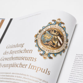 Das Bild zeigt ein aufgeschlagenes Buch mit einem detaillierten Emblem auf der linken Seite. Darunter befindet sich der Titel „Gründung des Bayerischen Gewerbemuseums als europäischer Impuls“.