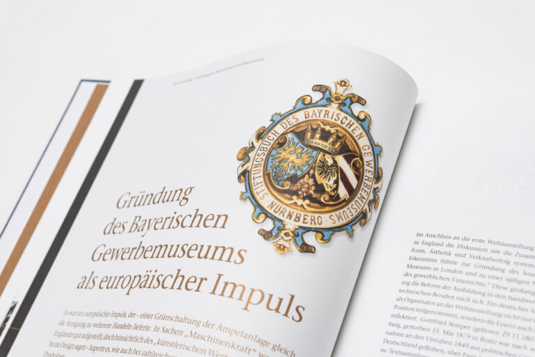 Das Bild zeigt ein aufgeschlagenes Buch mit einem detaillierten Emblem auf der linken Seite. Darunter befindet sich der Titel „Gründung des Bayerischen Gewerbemuseums als europäischer Impuls“.