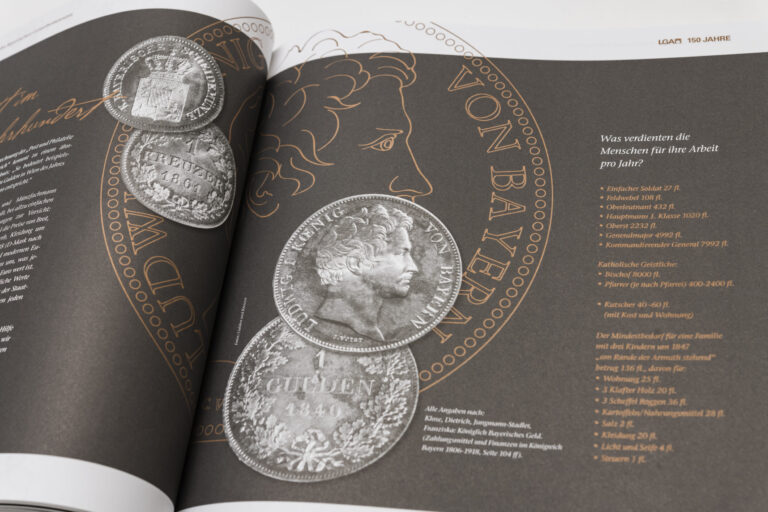 Das Bild zeigt ein aufgeschlagenes Buch mit dunklem Hintergrund und mehreren Abbildungen historischer Münzen. Auf der linken Seite sind zwei große Münzen mit feinen Gravuren zu sehen, auf der rechten Seite eine weitere Münze mit einem Profilporträt und der Aufschrift „VON BAYERN“. Daneben befindet sich ein Text mit historischen Gehaltsangaben verschiedener Berufe.