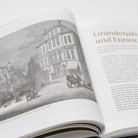 Das Bild zeigt ein aufgeschlagenes Buch mit einer Schwarz-Weiß-Zeichnung auf der linken Seite, die eine historische Straßenszene mit Gebäuden, Pferdekutschen und Menschen darstellt. Die Bildunterschrift verweist auf das Bayerische Gewerbemuseum. Auf der rechten Seite befindet sich ein Text mit dem Titel „Gründerjahre und Entwicklung“.