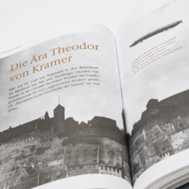 Das Bild zeigt ein aufgeschlagenes Buch mit einer Schwarz-Weiß-Fotografie einer Burg oder Festung auf einem Hügel auf der linken Seite. Daneben steht ein Text über die Ära Theodor von Kramer. Auf der rechten Seite ist ein Luftschiff (Zeppelin) abgebildet, begleitet von weiterem Text.
