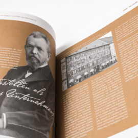 Das Bild zeigt ein geöffnetes Buch mit zwei sichtbaren Seiten. Auf der linken Seite ist ein schwarz-weißes Foto eines Mannes sowie ein deutscher Text zu sehen. Die rechte Seite zeigt ein weiteres schwarz-weißes Foto eines Gebäudes und ebenfalls deutschen Text.