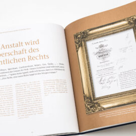 Das Bild zeigt ein geöffnetes Buch mit Text auf der linken Seite und einem gerahmten Dokument auf der rechten Seite. Die linke Seite enthält einen längeren deutschen Text mit erzählerischem Inhalt. Auf der rechten Seite ist ein offiziell wirkendes Dokument mit mehreren Unterschriften zu sehen.