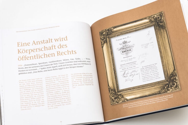 Das Bild zeigt ein geöffnetes Buch mit Text auf der linken Seite und einem gerahmten Dokument auf der rechten Seite. Die linke Seite enthält einen längeren deutschen Text mit erzählerischem Inhalt. Auf der rechten Seite ist ein offiziell wirkendes Dokument mit mehreren Unterschriften zu sehen.