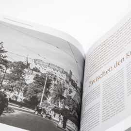 Das Bild zeigt ein geöffnetes Buch mit einer schwarz-weißen Fotografie auf der linken Seite und Text auf der rechten Seite. Die Fotografie zeigt eine Straßenszene mit Menschen, Bäumen, Gebäuden und Stromleitungen. Auf der rechten Seite steht ein großer deutscher Titel „Zwischen den Kriegen“ mit mehreren Absätzen Fließtext darunter.