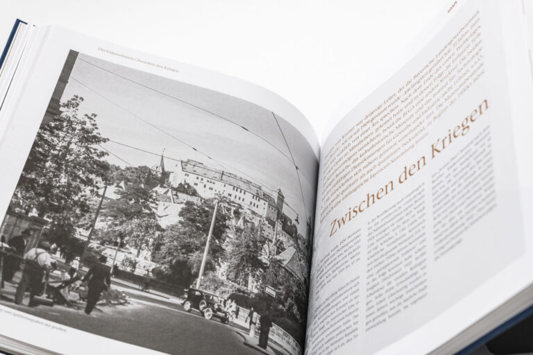 Das Bild zeigt ein geöffnetes Buch mit einer schwarz-weißen Fotografie auf der linken Seite und Text auf der rechten Seite. Die Fotografie zeigt eine Straßenszene mit Menschen, Bäumen, Gebäuden und Stromleitungen. Auf der rechten Seite steht ein großer deutscher Titel „Zwischen den Kriegen“ mit mehreren Absätzen Fließtext darunter.