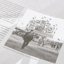 Das Bild zeigt eine Seite aus einem Buch oder Magazin mit deutschem Text und einer schwarz-weißen Fotografie. Die Fotografie zeigt das Volksfest von 1909 in der Fürther Straße in Nürnberg mit einem Mann im Vordergrund und einem großen Fahrgeschäft mit Fahnen im Hintergrund. Weitere Personen sind auf dem Festgelände unterwegs.