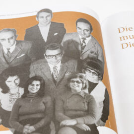 Das Bild zeigt eine Seite aus einem Buch oder Magazin mit einer alten schwarz-weißen Fotografie von acht Personen. Die Gruppe besteht aus fünf Männern und drei Frauen in formeller Kleidung, typisch für die Mitte des 20. Jahrhunderts. Der Hintergrund ist orange eingefärbt.