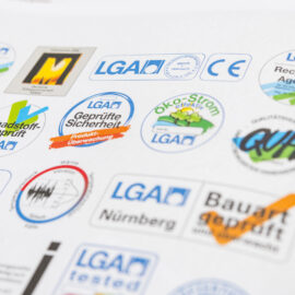 Das Bild zeigt eine Sammlung verschiedener Prüf- und Qualitätssiegel. Zu sehen sind unter anderem Symbole für Produktsicherheit, Schadstoffprüfung, Recycling, Ökostrom und CE-Kennzeichnung. Die Gestaltung umfasst unterschiedliche Farben und Formen der Siegel.