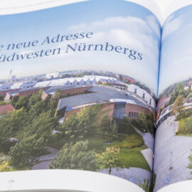 Das Bild zeigt ein geöffnetes Buch mit einer Luftaufnahme einer Stadtlandschaft. Zu sehen sind mehrere Gebäude, die von viel Grün umgeben sind, vermutlich im Südwesten Nürnbergs. Auf der linken Seite steht der Text „Eine neue Adresse im Südwesten Nürnbergs“.