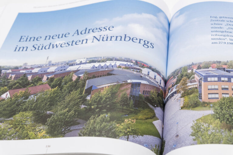 Das Bild zeigt ein geöffnetes Buch mit einer Luftaufnahme einer Stadtlandschaft. Zu sehen sind mehrere Gebäude, die von viel Grün umgeben sind, vermutlich im Südwesten Nürnbergs. Auf der linken Seite steht der Text „Eine neue Adresse im Südwesten Nürnbergs“.