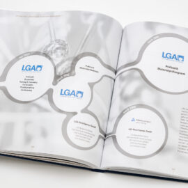 Das Bild zeigt ein geöffnetes Buch mit einem Diagramm, das sich über beide Seiten erstreckt. Das Diagramm besteht aus mehreren kreisförmigen Elementen, die durch Linien verbunden sind und jeweils Text sowie das LGA-Logo enthalten. Es stellt eine zeitliche Entwicklung von Organisationseinheiten vor und enthält Begriffe wie „vor 2002“, „nach 2002“, „Prüfstelle“, „Zertifizierung“ und „TÜV Rheinland Group“.