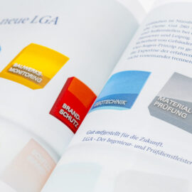 Das Bild zeigt ein geöffnetes Buch mit farbigen 3D-Blöcken auf der linken Seite, die verschiedene Kategorien darstellen. Die Blöcke sind mit Begriffen wie „PRÜFSTATIK“, „BAUWERKSMONITORING“, „BRANDSCHUTZ“, „GEOTECHNIK“ und „MATERIALPRÜFUNG“ beschriftet. Auf der rechten Seite befindet sich ein deutscher Textabschnitt mit einer Einleitung zur zukünftigen Ausrichtung der LGA im Bauwesen.