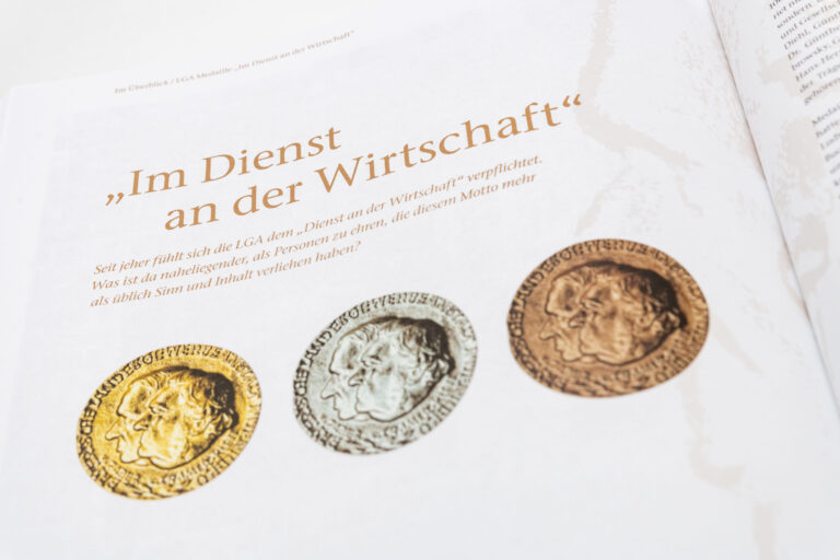 Das Bild zeigt eine Seite aus einer Publikation mit dem Titel „Im Dienst an der Wirtschaft“. Unter dem Titel steht ein erklärender Text zur Bedeutung dieses Mottos. Darunter sind drei Medaillen abgebildet – von links nach rechts in Gold, Silber und Bronze – jeweils mit zwei Profilporträts und umlaufender Beschriftung.