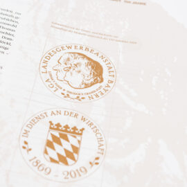 Das Bild zeigt eine Seite aus einer Publikation mit dem Titel „Im Dienst an der Wirtschaft“. Unter dem Titel steht ein erklärender Text zur Bedeutung dieses Mottos. Darunter sind drei Münzen abgebildet – von links nach rechts in Gold, Silber und Bronze – jeweils mit einem Profilporträt und umlaufender Beschriftung.