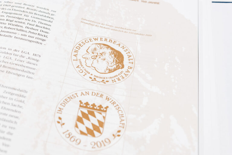 Das Bild zeigt eine Seite aus einer Publikation mit dem Titel „Im Dienst an der Wirtschaft“. Unter dem Titel steht ein erklärender Text zur Bedeutung dieses Mottos. Darunter sind drei Münzen abgebildet – von links nach rechts in Gold, Silber und Bronze – jeweils mit einem Profilporträt und umlaufender Beschriftung.