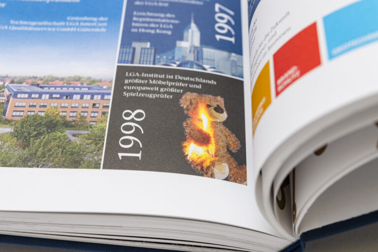 Das Bild zeigt ein geöffnetes Buch mit einer Zeitleiste, auf der die Jahre 1997 und 1998 hervorgehoben sind. Im Abschnitt zu 1998 steht, dass das LGA-Institut Deutschlands größter Möbelprüfer und europaweit größter Spielzeugprüfer ist. Daneben ist ein brennender Teddybär abgebildet.