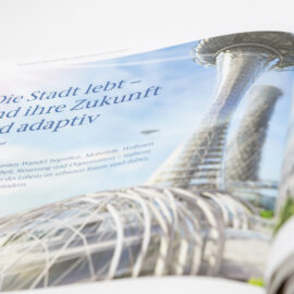 Das Bild zeigt ein geöffnetes Magazin mit einer futuristischen architektonischen Darstellung. Auf der Seite steht ein Text mit dem Titel „Die Stadt lebt – und ihre Zukunft wird adaptiv“. Der Text beschreibt den schnellen Wandel städtischen Lebens in Bereichen wie Mobilität, Wohnen, Arbeiten, Sicherheit und Organisation.
