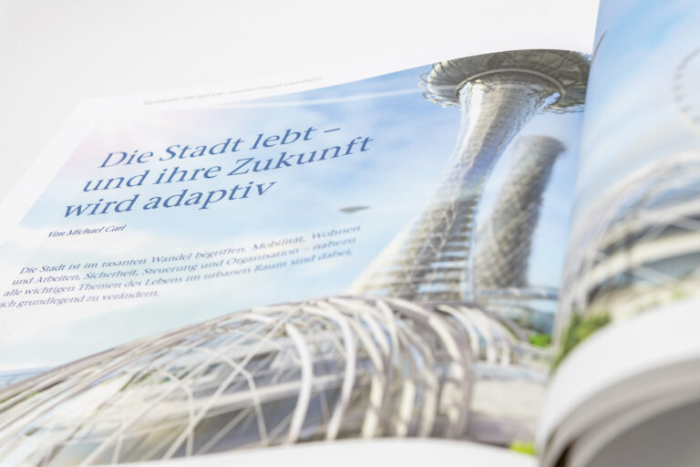 Das Bild zeigt ein geöffnetes Magazin mit einer futuristischen architektonischen Darstellung. Auf der Seite steht ein Text mit dem Titel „Die Stadt lebt – und ihre Zukunft wird adaptiv“. Der Text beschreibt den schnellen Wandel städtischen Lebens in Bereichen wie Mobilität, Wohnen, Arbeiten, Sicherheit und Organisation.